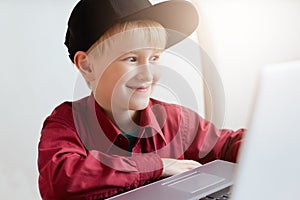 A happy little boy in trendy clothes relaxing during lunch at modern cafe, sitting in front of open laptop having surprised expres