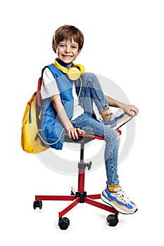 Happy little boy wearing headphone and yellow backpack sitting on red chair on white background. Back to School.