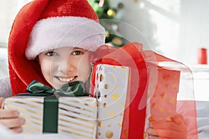 Happy little boy wearing a Santa hat looking at camera holding many gifts boxes at morning at home. Merry Christmas concept