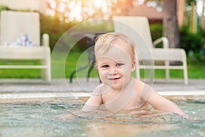 Happy little caucasian blond toddler boy swimming in wading pool on bright summer day at resort. Adorable baby enjoying