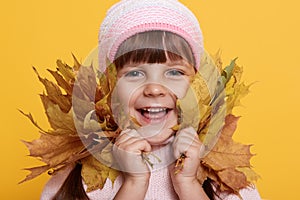 Happy little child, baby girl laughing and playing with autumn leaves, looks at camera with charming smile, wearing pink cap,