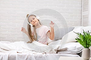 Little girl listening to the music with the headphones on bed.