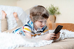 Happy little child boy playing online game, watching video on cellphone, lying on sofa entertaining in living room