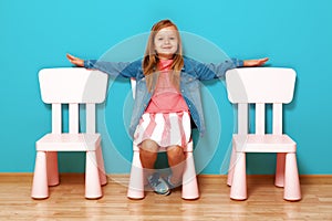 A happy little child girl with arms extended to the side sits on a chair against the background of a blue wall.