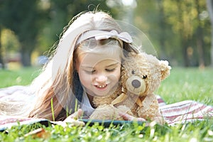 Happy little child girl looking in her mobile phone with her favorite teddy bear toy outdoors in summer park