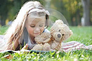 Happy little child girl playing her sellphone together with favorite teddy bear toy outdoors in summer park