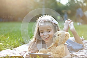 Happy little child girl playing her sellphone together with favorite teddy bear toy outdoors in summer park