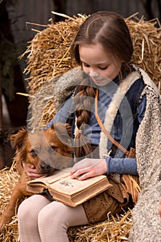 Happy little child girl reads old book with dog on hay