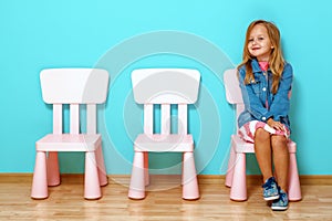 Happy little child  girl is sitting on the chair against the background of the blue wall. Nearby are empty chairs.