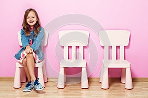 Happy little child  girl is sitting on the chair against the background of the pink wall. Nearby are empty chairs.