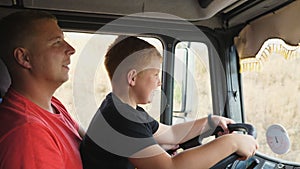 Happy little child holds his hands on steering wheel sitting on lap of daddy and learning to drive the truck. Lorry