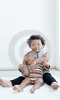 Happy Little cute African chubby kid girl hugging her newborn baby sister in arms while sitting playing together on floor at home