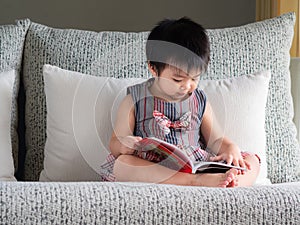 Happy little cute girl is reading the book on the white sofa. Ed