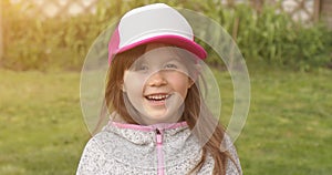 Happy little cute girl in trucker cap smiling and looking at camera while standing in summer garden