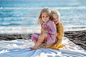 Happy little cute girls enjoying sunny day at the beach, standing on the coast and looking at the waves. Family summer vacation.