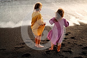 Happy little cute girls enjoying sunny day at the beach, standing on the coast and looking at the waves. Family summer vacation.