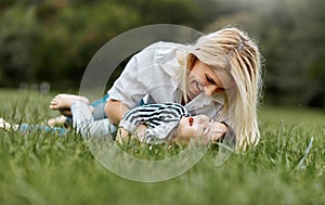 Happy little daughter lying on the green grass and playing with her smiling mother in the park. Loving woman and her little kid