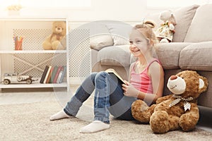 Happy little female child and her teddy bear reading book on the floor at home