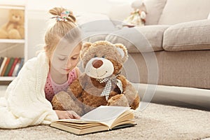 Happy little female child and her teddy bear reading book on the floor at home