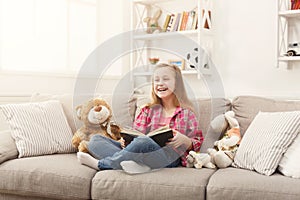 Happy little female child hugging her teddy bear and reading book on sofa at home