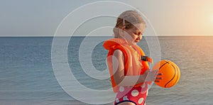 Happy little girl with blonde hair in kids swim vest with inflatable orange ball in hands, colorful seascape sunset