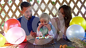 Happy little girl blowing out candle on her birthday cake and her loving parents kiss the her and clap the hands