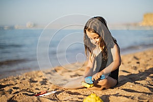 Happy little girl building sandcastle with plastic shovel, sitting on wet sand by water.