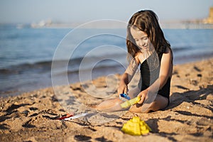 Happy little girl building sandcastle with plastic shovel, sitting on wet sand by water.