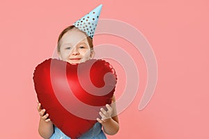 Happy little girl in a cap holds a big heart shaped balloon. Portrait of a child on a pink background. Copy space. Closeup