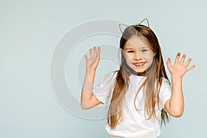 Happy little girl in cat ears raises her hands up on a blue background with copy space.