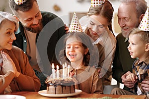 Happy little girl celebrating birthday with family at home, closing   eyes and  making wish with cake with lit candles