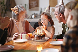 Happy little girl celebrating birthday with family at home, looking at cake with lit candles