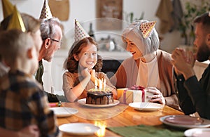 Happy little girl celebrating birthday with family at home, looking at cake with lit candles