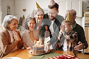 Happy little girl celebrating birthday with family at home, looking at cake with lit candles