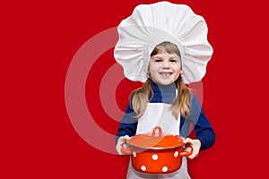 Happy little girl in chef uniform holds saucepan isolated on red. Kid chef