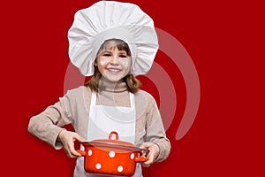 Happy little girl in chef uniform holds saucepan isolated on red. Kid chef