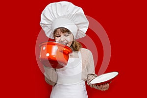 Happy little girl in chef uniform holds saucepan isolated on red. Kid chef