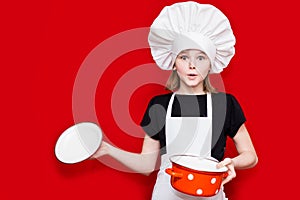 Happy little girl in chef uniform holds saucepan isolated on red. Kid chef. Cooking Concept