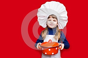 Happy little girl in chef uniform holds saucepan on red. Kid chef. Cooking Concept