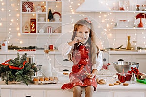 A happy little girl in the Christmas kitchen is sitting on the table with cookies in her hands and eating it