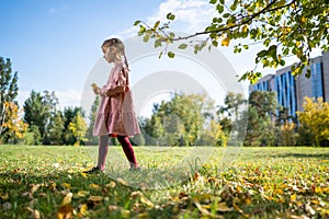 Happy little girl collects and plays with autumn fallen leaves in autumn park. Fall season