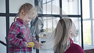 Happy little girl combing blond Caucasian woman with toy hairbrush. Patient young mother spending weekends with active