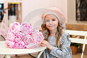 Happy little girl with curly hair in a dress and in a beret sits at a table with a bouquet of pink peonies in a street vintage caf