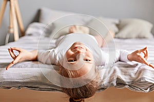 Happy little girl with dark hair lying upside down on bed at home, kid playing having fun showing victory looking smiling to