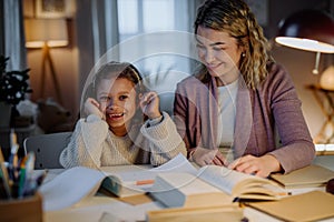 Happy little girl doing homework with her mother and looking at camera in evening at home.