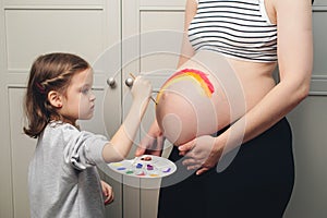 Happy little girl drawing rainbow on pregnant belly her mother. Baby birth expecting time and belly painting