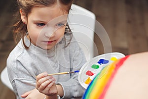 Happy little girl drawing rainbow on pregnant belly her mother. Baby birth expecting time and belly painting