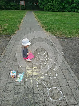 Happy little girl draws with colored crayons sitting on the sidewalk. Children draw with chalk on the street in the Park on the ro