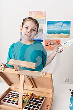 Happy little girl with easel, palette and brush. The young artist dreams of a vacation at the seaside.