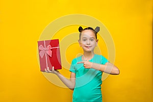Happy little girl with funny tails standing isolated over yellow background holding red gift box. Looking camera